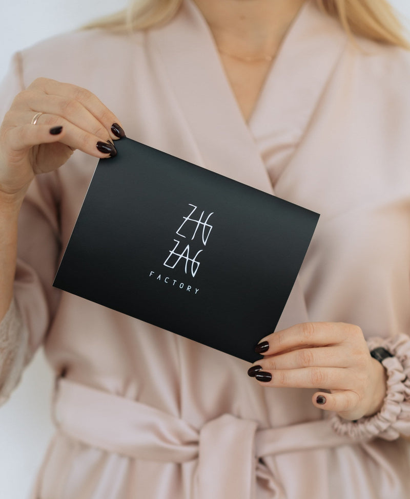 Person holding a black box with 'Zig Zag Factory' Gift Card, wearing a beige robe.